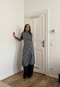 Femme aux longs cheveux noirs portant des lunettes de soleil, un col roulé gris, une jupe à carreaux par-dessus un pantalon noir et des chaussures noires, debout à l'intérieur près d'une porte blanche.