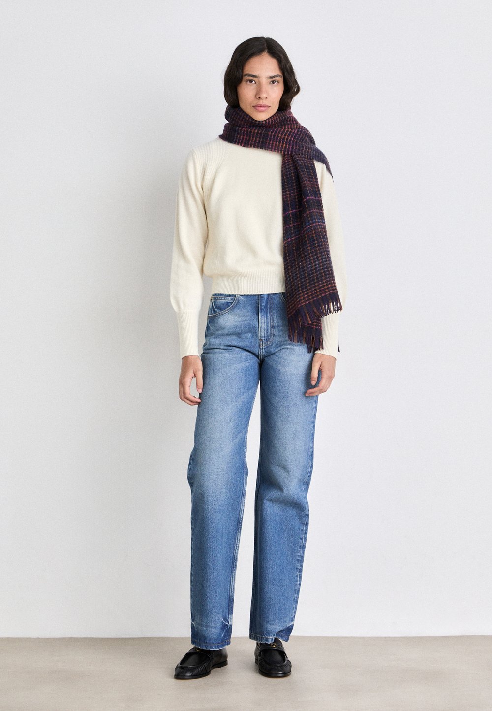 Crema maglione lavorato a maglia con orlo ampio a coste, abbinato a jeans dritti di un blu chiaro. Una sciarpa a quadri scuri pende attorno al collo. Scarpe nere.