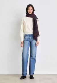 Crema maglione lavorato a maglia con orlo ampio a coste, abbinato a jeans dritti di un blu chiaro. Una sciarpa a quadri scuri pende attorno al collo. Scarpe nere.