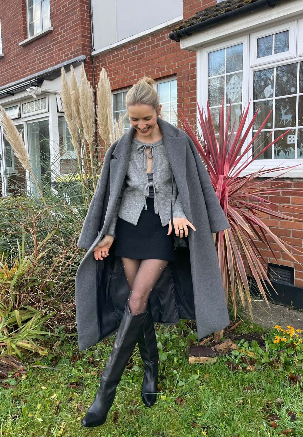 Γκρίζα υπερμεγέθη παλτό, υφασμάτινη γκρι μπλούζα με φιόγκo, μαύρη μίνι φούστα, καλσόν με πουά και μαύρες μπ ότες μέχρι το γόνατο μπροστά σε φόντο κήπου.
