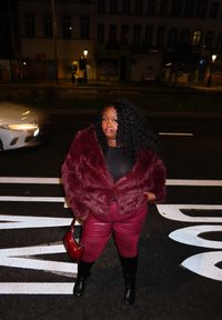 Γυναίκα με κατσαρά μαλλιά, φοράει ένα μπορντό μπουφάν και παντελόνι από ψεύτικη γούνα, κρατώντας μια κόκκινη τσάντα, στέκεται σε διάβαση πεζών σε νυχτερινό δρόμο.