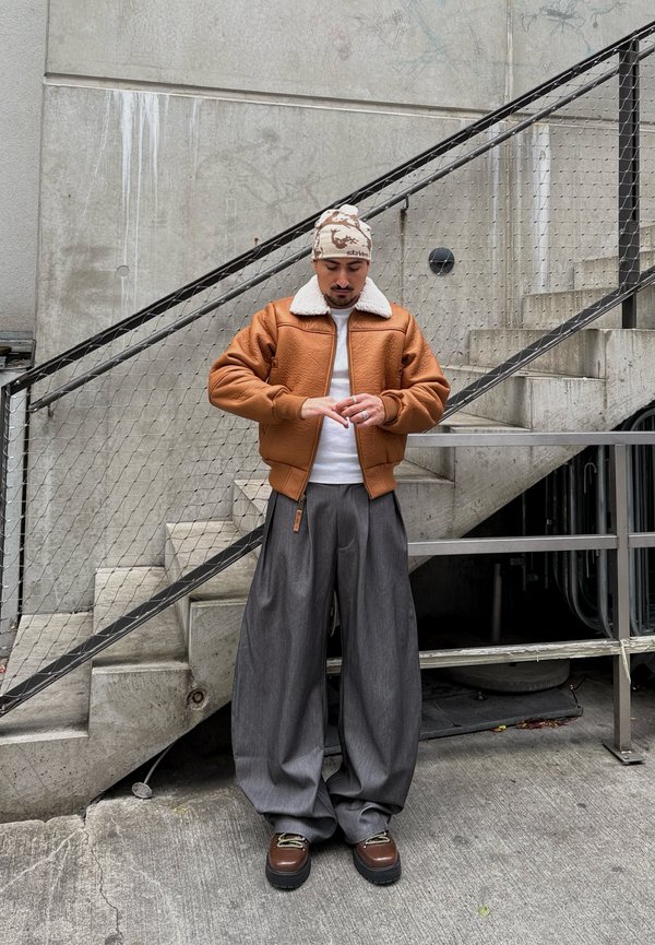 Brown bomber jacket with a white fur collar, paired with loose grey trousers and brown platform shoes. Background features concrete steps and a railing.