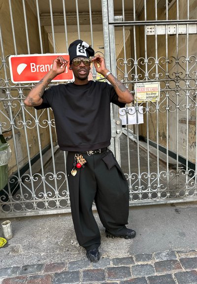 Camiseta negra corta y pantalones negros anchos, complementados con un cinturón de diseño y un llavero. Lentes de sol oscuros y detalle de sombrero único visible.