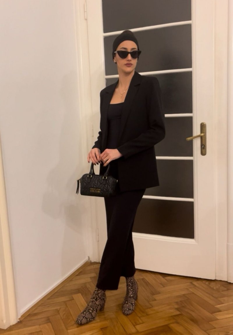 Femme debout à l'intérieur portant un blazer noir et un pantalon, des lunettes de soleil foncées, un foulard sur la tête, tenant un sac à main de créateur noir, et des bottines à imprimé serpent.