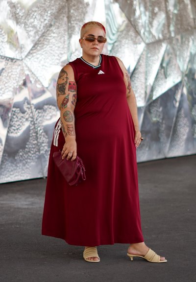 Vestido sin mangas de color burdeos con la marca Adidas. El modelo sostiene un bolso de mano burdeos, combinado con sandalias de lona beige. Decorado con tatuajes.