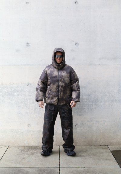 Chaqueta de plumas en un patrón de tie-dye oscuro, con cremallera y capucha. Combinada con pantalones cargo negros holgados y zapatos gruesos negros.