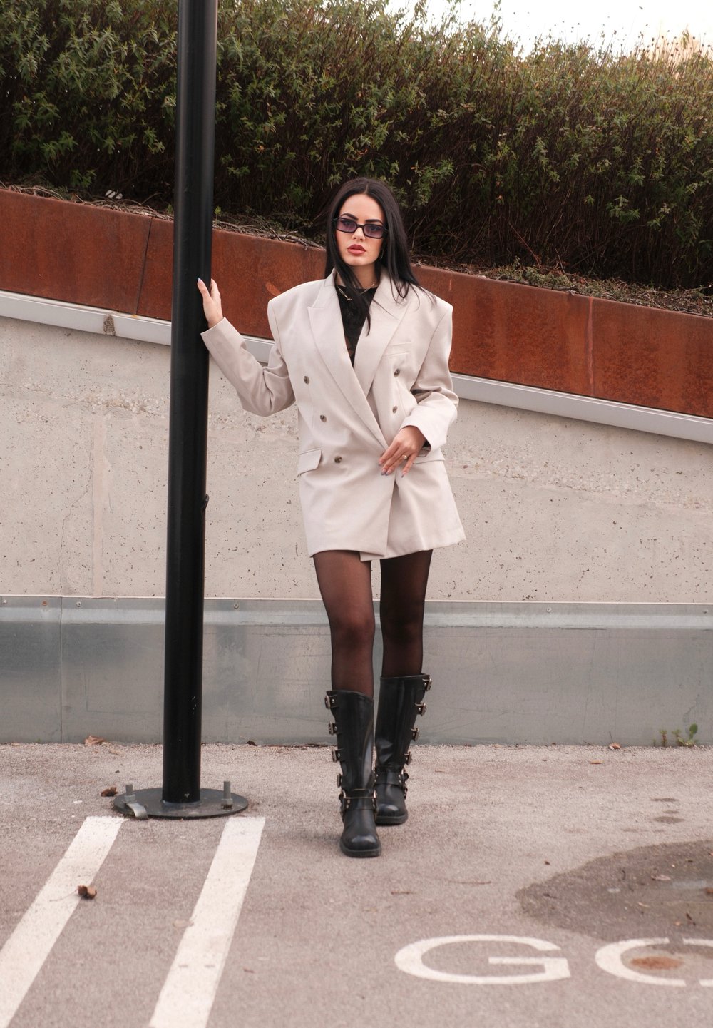 Μπεζ διπλόχαρο φόρεμα σακάκι με φαρδιές ώμους, συνδυασμένο με μαύρες μπότες μέχρι το μηρό. Διάφανες μαύρες καλσόν και παραλληλόγραμμα γυαλιά ηλίου.