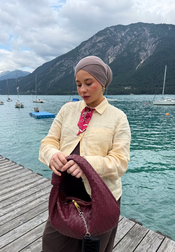 Femme portant une veste beige et un foulard sur la tête se tient sur un ponton en bois au bord d'un lac avec des montagnes et des voiliers en arrière-plan, tenant un sac bordeaux.
