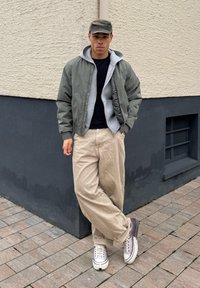 Jeune personne portant une veste verte, un sweat à capuche gris, un pantalon beige et des baskets blanches, appuyée contre un mur extérieur bicolore sur un trottoir pavé.