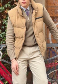 Vaaleanruskean neuleen päällä tan-värinen toppiliivi, jossa vetoketjullinen tasku, yhdistetty kermaan sävyisiin housuihin. Taustalla vihreää kasvillisuutta ja punainen polkupyörä.