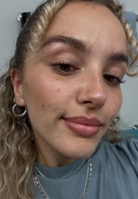 Selvportrett med bølgete, skulderlangt hår. Personen har på seg sølvfargede hoops, en grå skjorte, og har en lys leppestift.