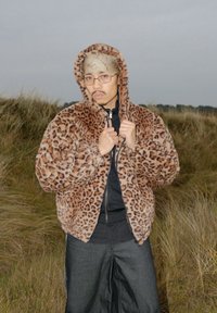 Veste zippée en fausse fourrure à imprimé léopard marron avec capuche, portée sur un haut noir. Le modèle se tient dans une herbe haute, tenant la veste fermée.