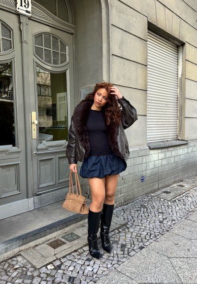 Joven mujer con chaqueta de cuero negra con cuello de piel, falda azul marino y botas negras hasta la rodilla, de pie sobre una acera adoquinada frente a un edificio gris.