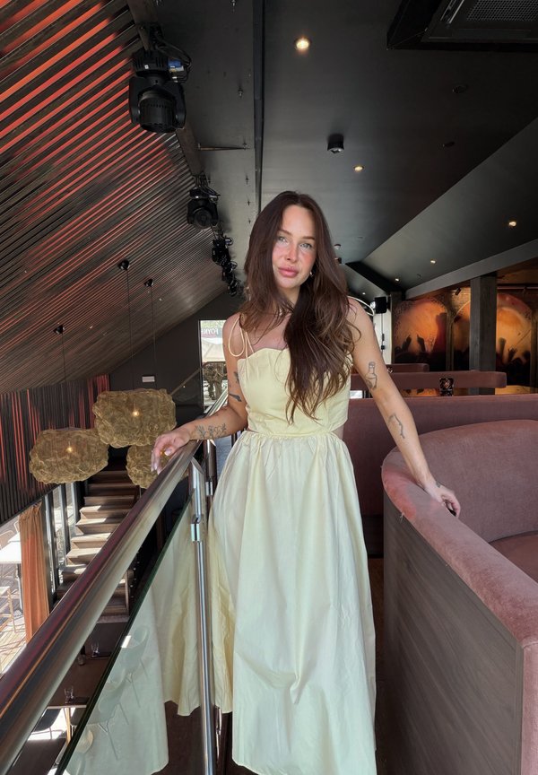 Jonge vrouw met lang bruin haar en tatoeages draagt een crème jurk en leunt op een glazen reling binnen een modern, schemerig verlicht restaurant of lounge.