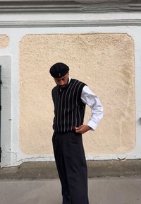 Svart og hvit vertikalt stripete genser vest over en hvit skjorte, kombinert med svarte bukser. Bakgrunnen har en teksturert beige vegg.