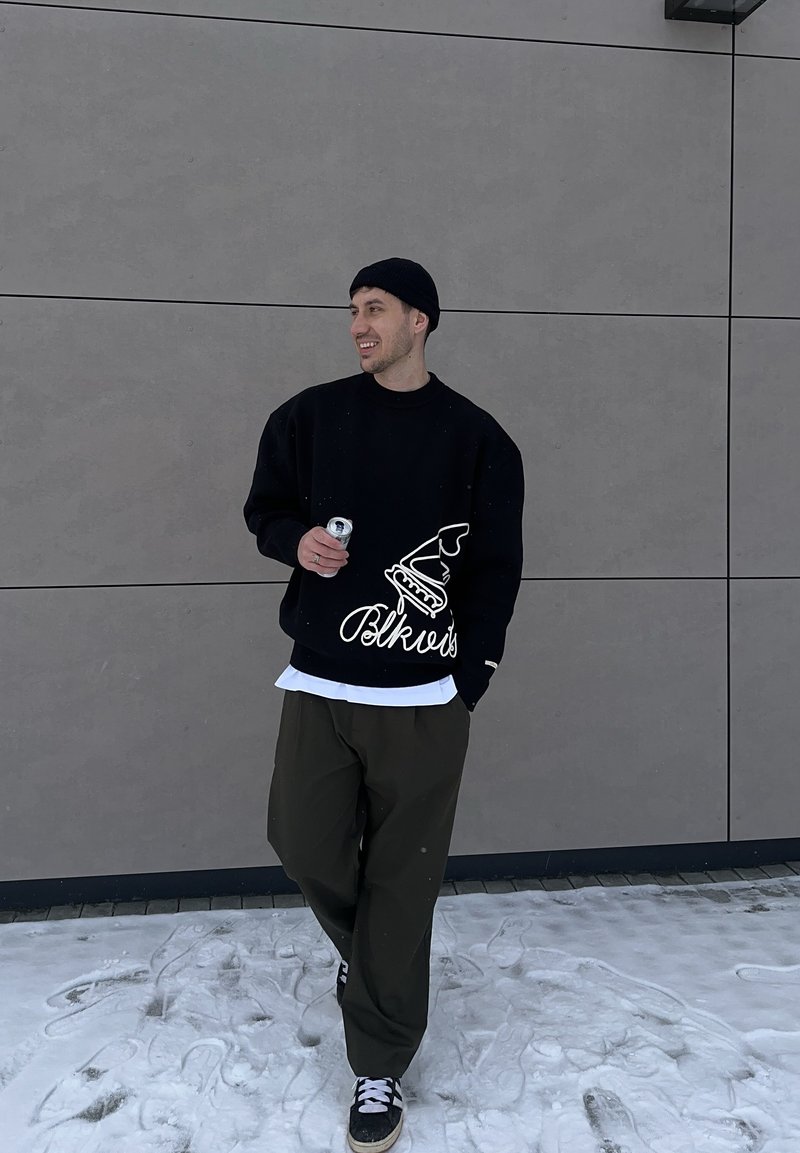 Homme portant un bonnet noir, un sweat-shirt noir surdimensionné avec un motif blanc, un pantalon olive et des baskets, tenant une canette en se tenant sur un sol enneigé devant un mur gris.