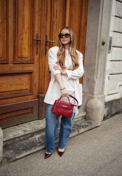 Mujer con cabello largo y rubio, vestida con blazer blanco, vaqueros azules, zapatos burdeos, gafas de sol y sosteniendo un bolso rojo, de pie junto a una puerta de madera.