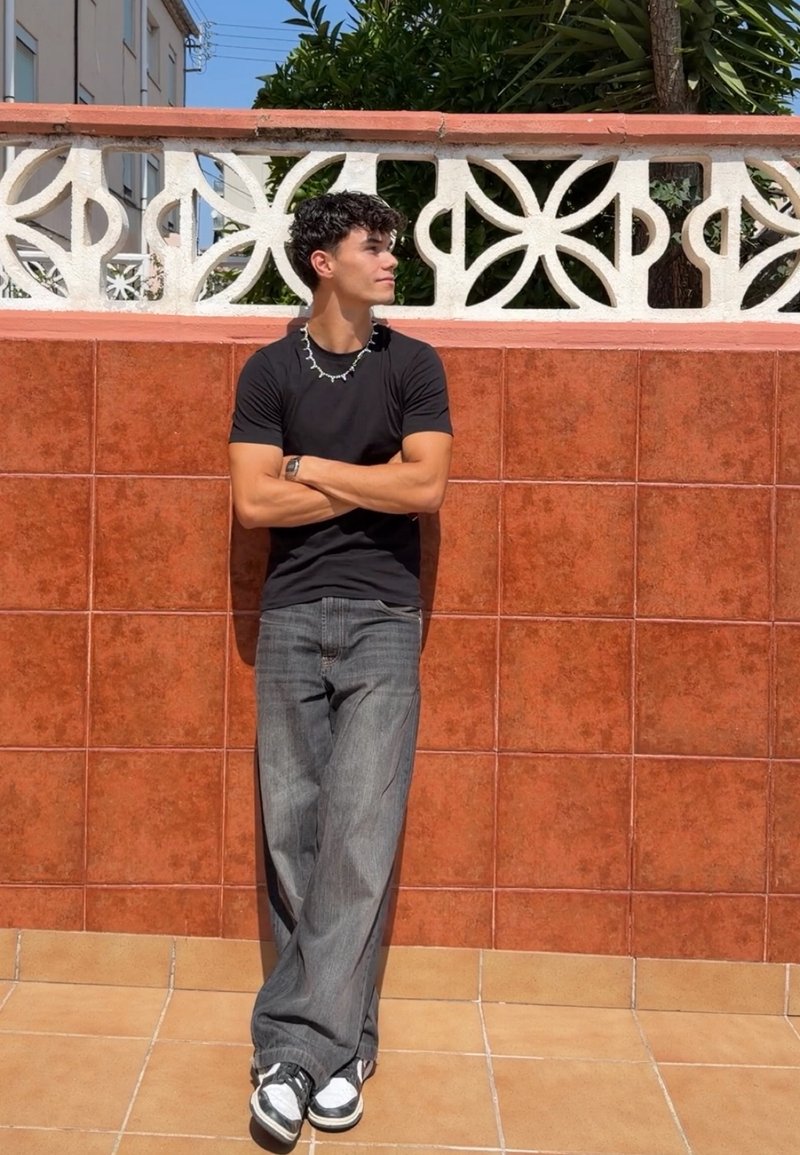 Jeune homme en t-shirt noir et jean gris appuyé contre un mur carrelé rouge, les bras croisés, regardant sur le côté dans un cadre extérieur ensoleillé.