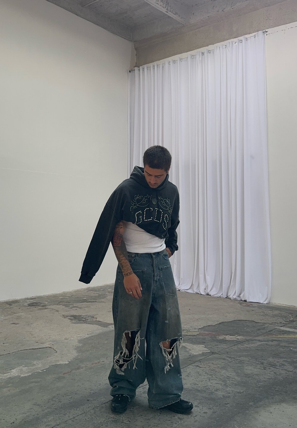 Black cropped hoodie featuring a graphic design, worn over a white shirt. Baggy distressed blue jeans with large rips. Black footwear. Plain background.