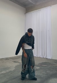 Black cropped hoodie featuring a graphic design, worn over a white shirt. Baggy distressed blue jeans with large rips. Black footwear. Plain background.