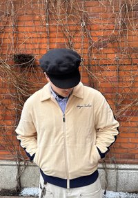 Homme portant une veste beige zippée et une casquette noire, les mains dans les poches, se tenant devant un mur en briques couvert de vignes sèches.