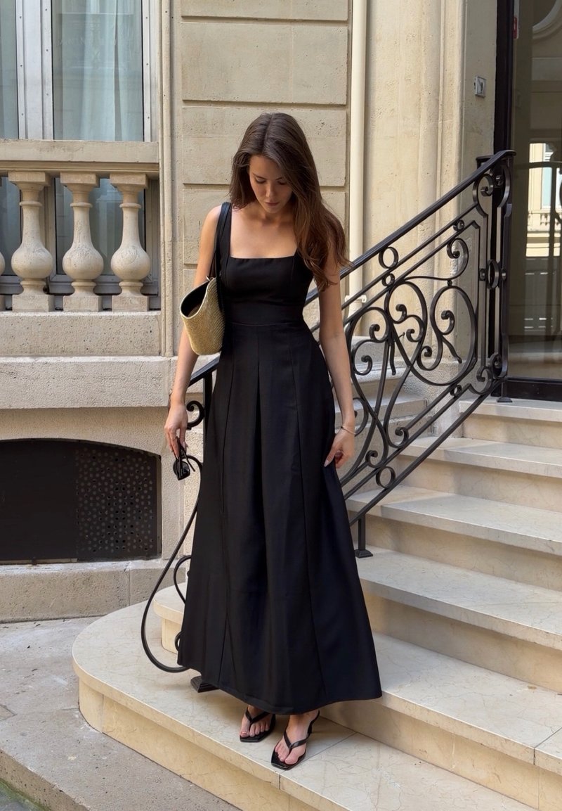 Femme en longue robe noire debout sur des marches en pierre, tenant des lunettes de soleil et un sac bandoulière tressé, regardant vers le bas, près d'une rampe ornée.