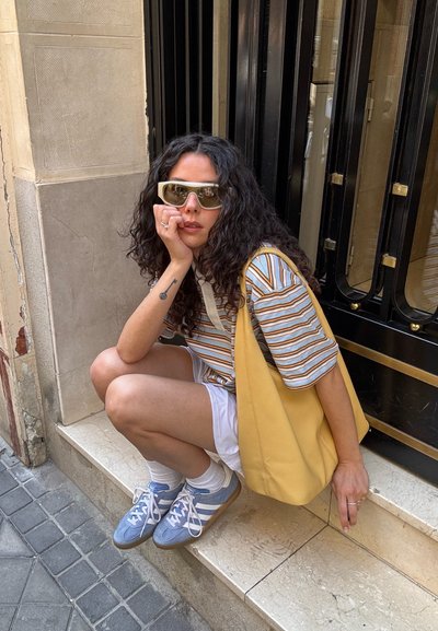Top de manga corta a rayas en tonos neutros, bolsa amarilla, pantalones cortos blancos, zapatillas deportivas azules y gafas de sol. Cabello rizado, sentado en escalones de piedra.
