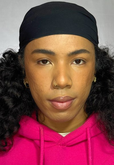 Persona con cabello rizado que lleva un pañuelo negro en la cabeza y una sudadera con capucha de color rosa brillante. El rostro muestra una complexión natural con un piercing en la nariz y maquillaje sutil.