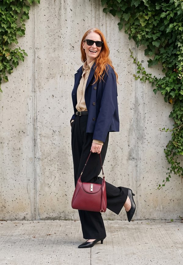 Vrouw met lang rood haar die een zonnebril draagt, een marineblauw jasje, een beige hemd, zwarte broek en hakken, houdt een rode handtas vast en staat bij een betonnen muur met klimop.