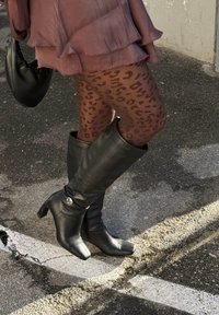 Bottes en cuir noires montant jusqu'aux genoux avec un bout carré, dotées d'un talon haut fin et d'un petit accent argenté. Portées avec des collants imprimés léopard marron.