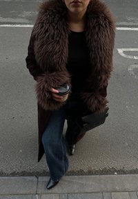 Casaco de pêlo sintético castanho com punhos combinantes, usado sobre um top preto. Jeans de perna larga azuis e sapatos de bico fino pretos. Segura uma chávena de café.