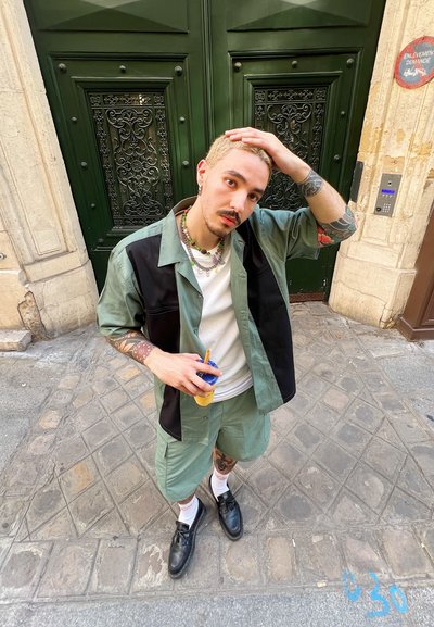 Camisa de manga corta verde claro y negra sobre una camisa blanca, pantalones cortos a juego, zapatos negros y calcetines blancos. Sosteniendo una bebida.