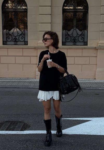 Camiseta negra de gran tamaño, falda blanca con volantes, botas negras hasta la rodilla. Lleva un bolso negro y una taza con una pajita.