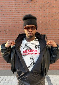 Fekete felfújt dzseki cipzáras elülső résszel, alatta fehér grafikás póló fekete rajzokkal és piros felirattal. Barna napszemüveg és fekete sapka.