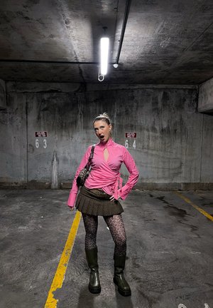Mujer joven con top rosa, falda marrón, leggings con estampado de leopardo y botas, posando en un estacionamiento de concreto con poca iluminación cerca de los espacios 63 y 64.