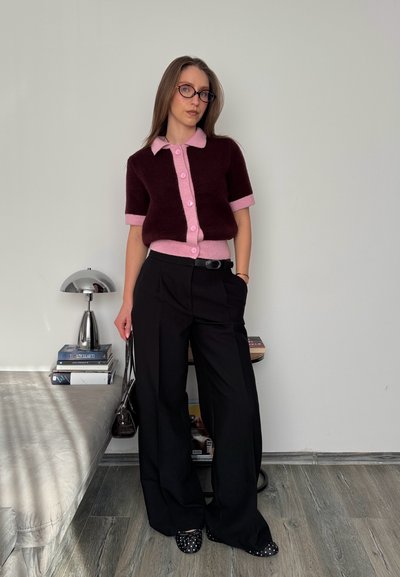 Mujer con suéter de manga corta en tonos morado y rosa con botones, pantalones negros de pierna ancha, gafas, sosteniendo una bolsa pequeña, de pie en una habitación minimalista con libros y lámpara.