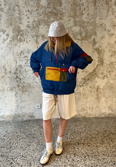 Chaqueta de pullover azul con bolsillos amarillos y verdes, detalles rojos y dobladillo con cordón; llevada sobre pantalones cortos blancos y combinada con un sombrero tipo cubo blanco.