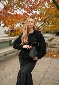 Pulover negru, texturat, cu detalii asemănătoare cu penele, asortat cu o fustă neagră cu tiv cu volane. Ține în mână o geantă mică neagră.