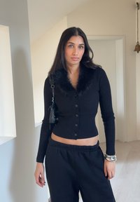 Chaqueta negra de punto con cuello de piel sintética, con botones en el frente. Combinada con pantalones negros de corte holgado y pulseras plateadas en la muñeca.