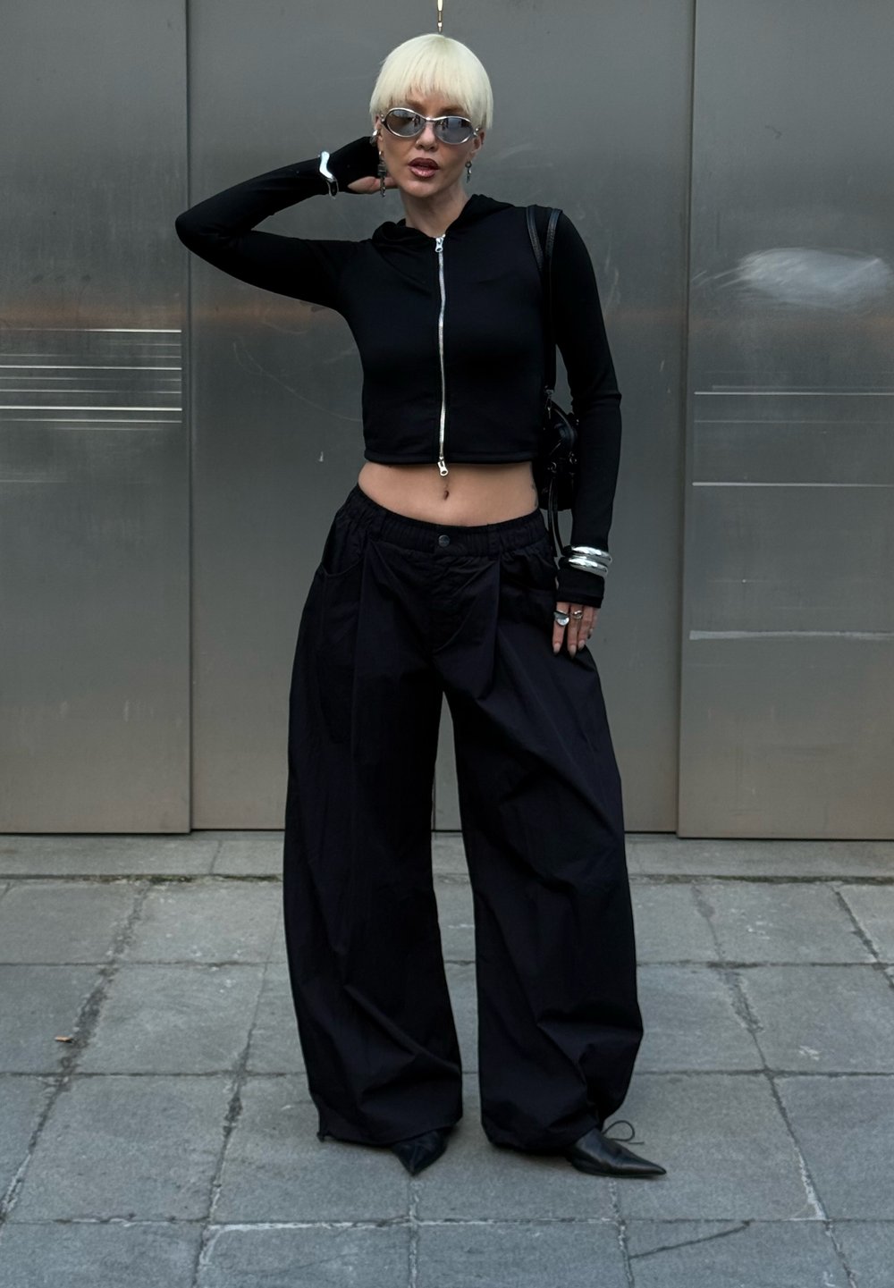 Czarna krótka bluza zapinana na zamek, oversize'owe czarne spodnie z kieszeniami, srebrne akcesoria i okulary przeciwsłoneczne. Gładki materiał, luźny krój, minimalistyczny design.
