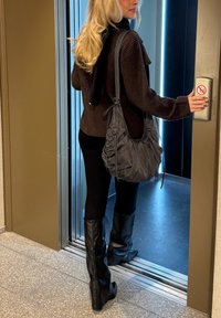 Fekete térdig érő, testhezálló csizmák, fekete leggings, sötét bordázott pulóver és válltáska. A személy egy lift közelében áll.