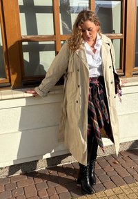 Šviesiai ruda trenčkotė ant baltos bluzos, sluoksniuota raudona ir žalia langeliais sijonu, derinama su juodais kelių ilgio botais, stovinti prie lango.