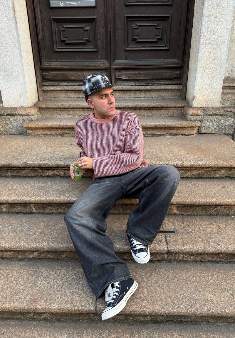 Casual outfit featuring a striped red and white sweater and loose black denim jeans. Black sneakers and a patterned cap. Green drink in hand.