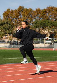 Atleta a correr com um casaco preto e leggings, em plena passada numa pista de corrida, usando sapatos desportivos brancos e cinzentos. Relva verde ao fundo.