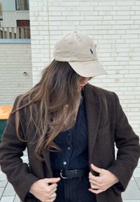 Jeune personne aux cheveux longs portant une casquette beige, un manteau brun foncé, un cardigan en maille bleu marine et un pantalon noir, ajustant son manteau à l'extérieur.