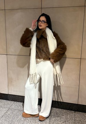 Chaqueta de piel sintética marrón, bufanda de punto blanca con flecos, pantalones anchos blancos y calzado marrón. Modelo posando contra una pared clara.