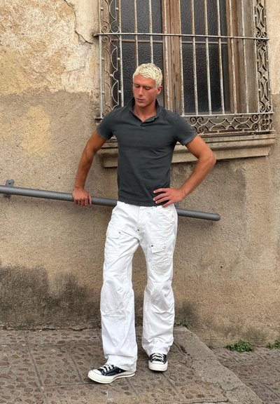 Modelo vistiendo una camiseta polo negra, pantalones cargo blancos con bolsillos y zapatos de lona negros, apoyado contra un poste gris frente a una pared texturizada.