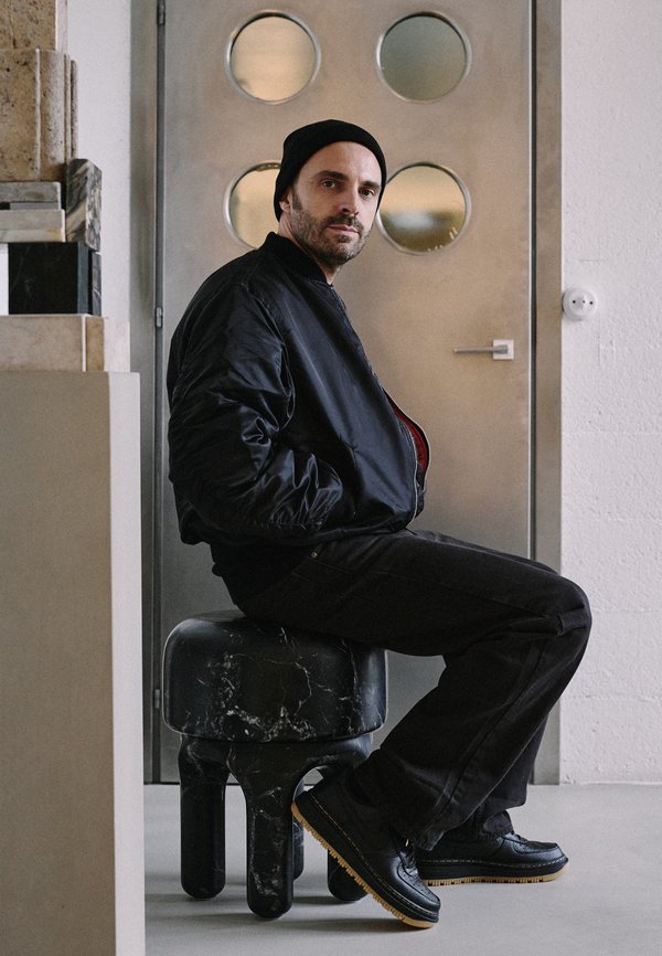 Giacca bomber nera, pantaloni neri e scarpe nere con suole gialle. Seduto su uno sgabello rotondo in marmo nero in un interno moderno.