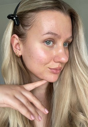 Cheveux blonds avec une pince noire, mettant en valeur une peau douce. Des boucles d'oreilles en or ajoutent du détail. Vernis à ongles rose clair sur les doigts. Éclairage naturel.