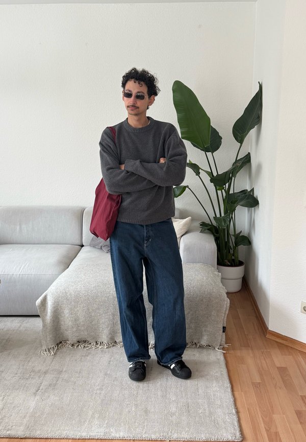 Mann med krøllete hår og solbriller står med armene i kors, iført grå genser, vide jeans, svarte joggesko og bærer en rød veske i stuen.